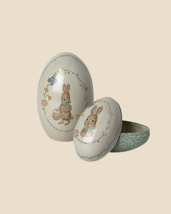 The Maileg Easter Egg set includes two green, oval boxes with vintage-style rabbit illustrations; one is closed and the other open, displaying intricate designs reminiscent of Dorthe Mailil embroidery. The plain background enhances their charm. Ships in approximately one week.