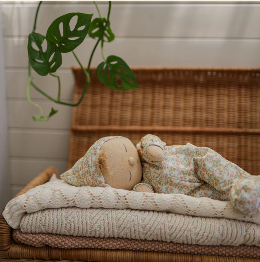 An Olli Ella Dozy Dinkums - Custard - Garden Floral doll in floral pajamas and cap rests on folded knitted blankets. A wicker chest and potted plant with vibrant leaves complete the background, making it an ideal Easter companion.