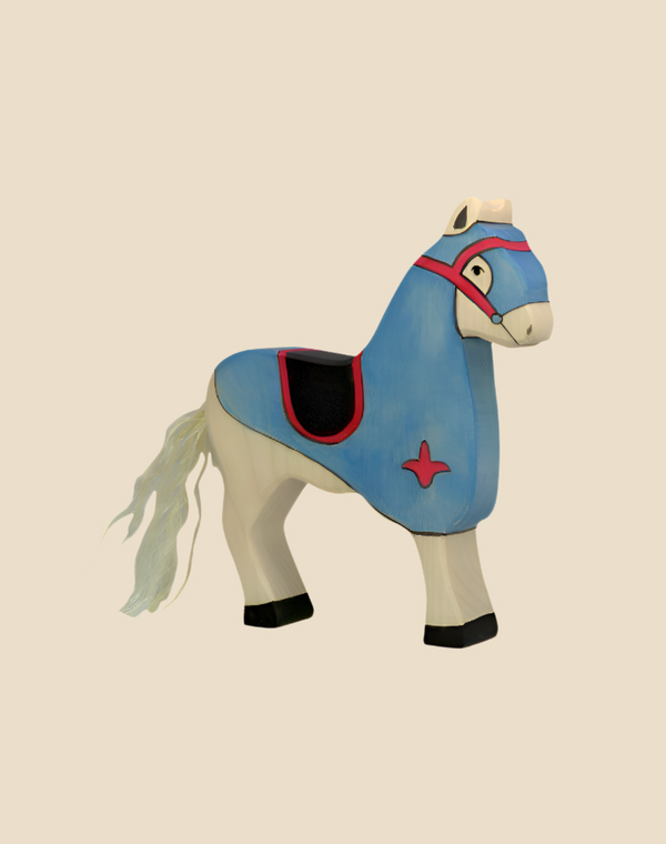 The Holztiger Tournament Horse, Blue, is a handcrafted wood toy featuring blue and white paint, black hooves, a pink and black saddle, and a long white tail. Ships in one week and adds European charm to playtime. Set against a beige background.
