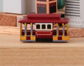The San Francisco Music Box - Bus Replacement, a small toy streetcar with a red roof and yellow sides, sits on a wooden surface with miniature buildings blurred in the background.