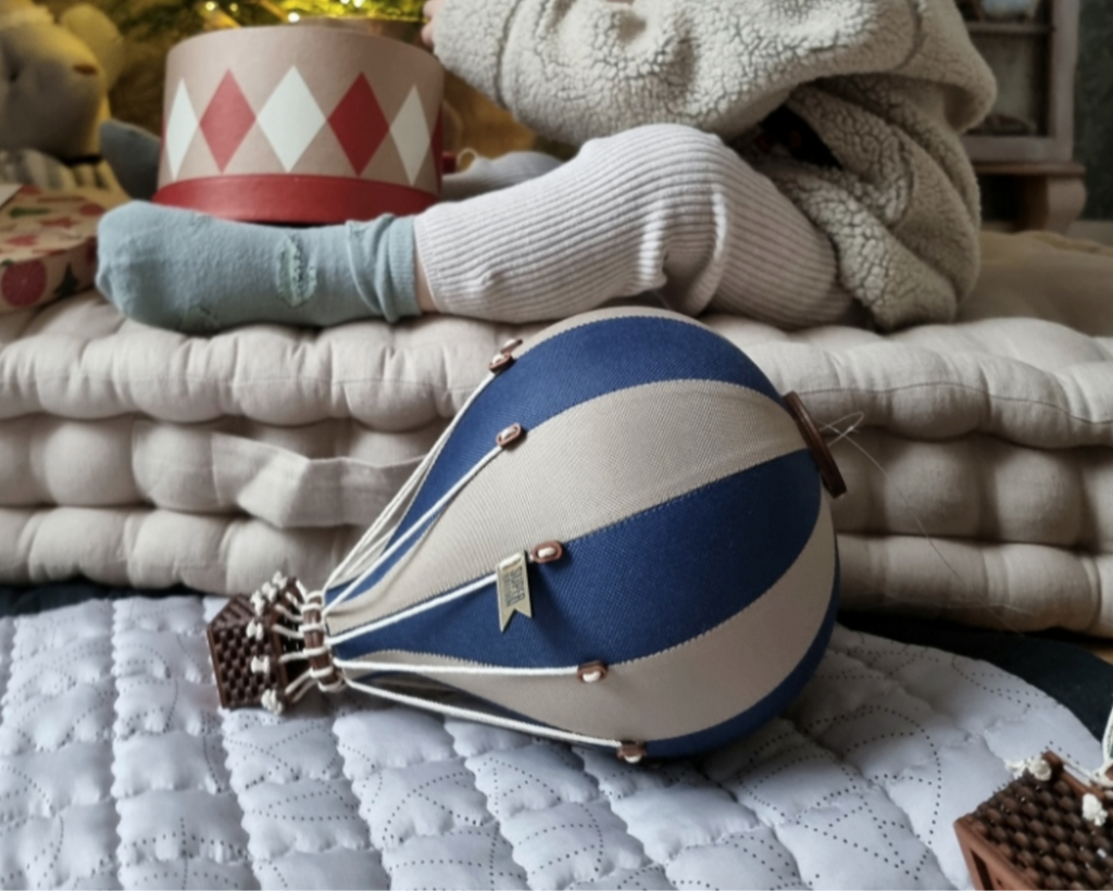 A light brown and navy blue handmade hot air balloon toy rests on a quilted mat, with folded blankets and a hat in the background. The softly lit scene creates a cozy atmosphere, perfect for adding charm to any children's room décor.
