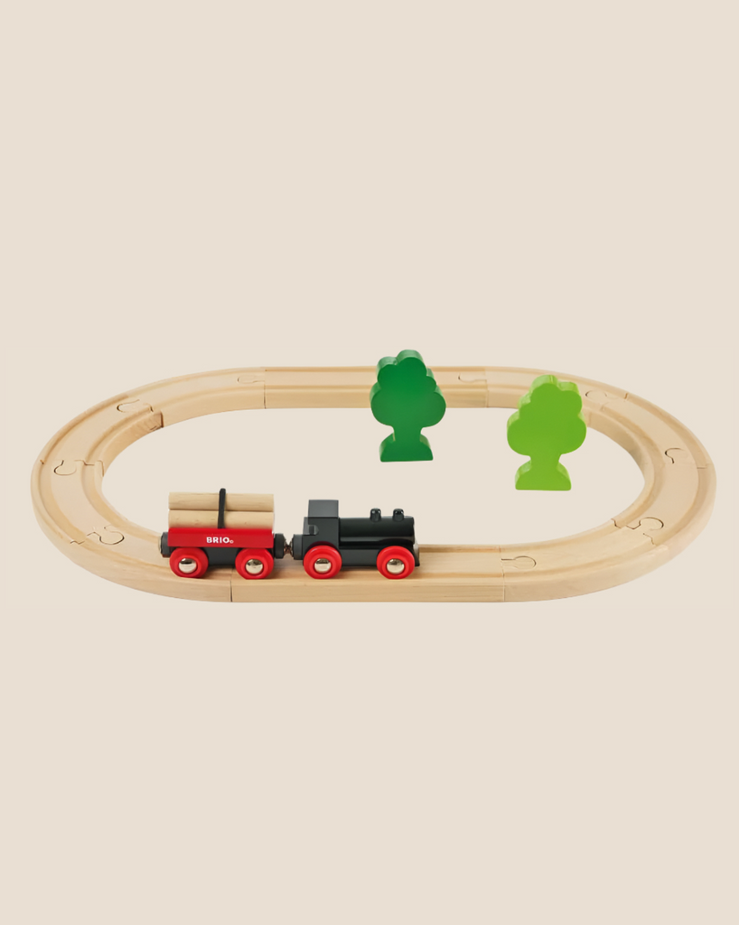 The BRIO Little Forest Train Set features a classic wooden oval track, a black and red train carrying logs, and two green trees—an ideal starter set for imaginative train play.