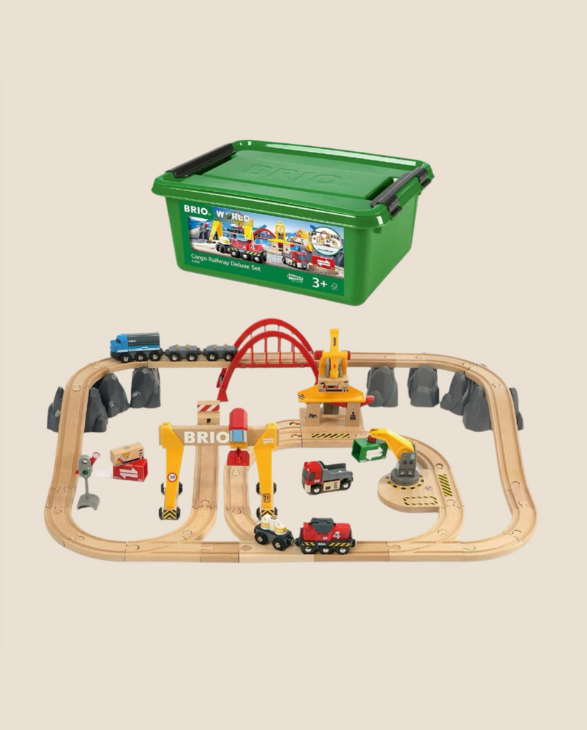 The BRIO Cargo Railway Deluxe S train set with tracks, trains, cranes, trucks, and accessories is displayed on a light surface. Above it floats a green storage box showing the BRIO logo and product images.