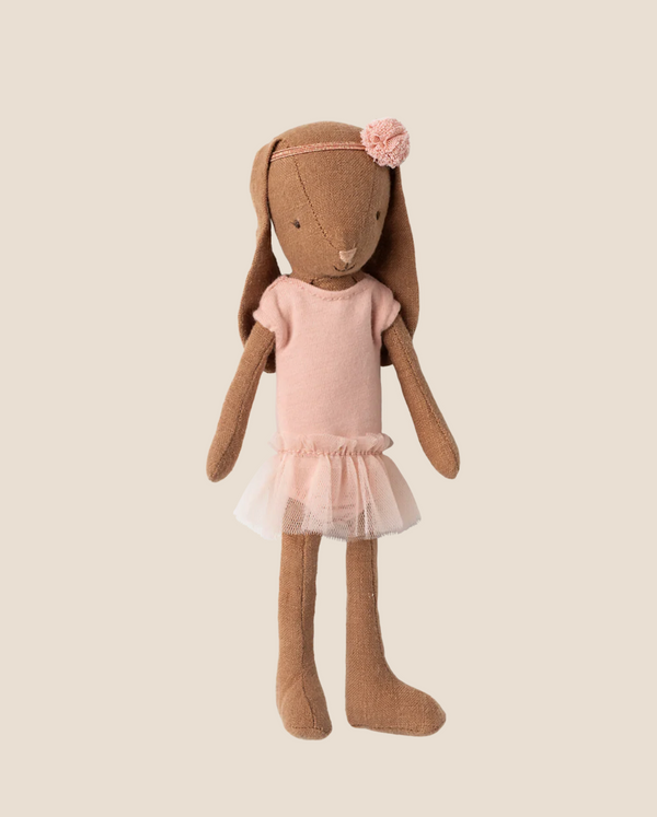 A Maileg Bunny, Size 1, in chocolate brown, wears a rose ballet suit and skirt. This adorable doll resembles a tiny ballerina and stands out beautifully against a plain beige background.