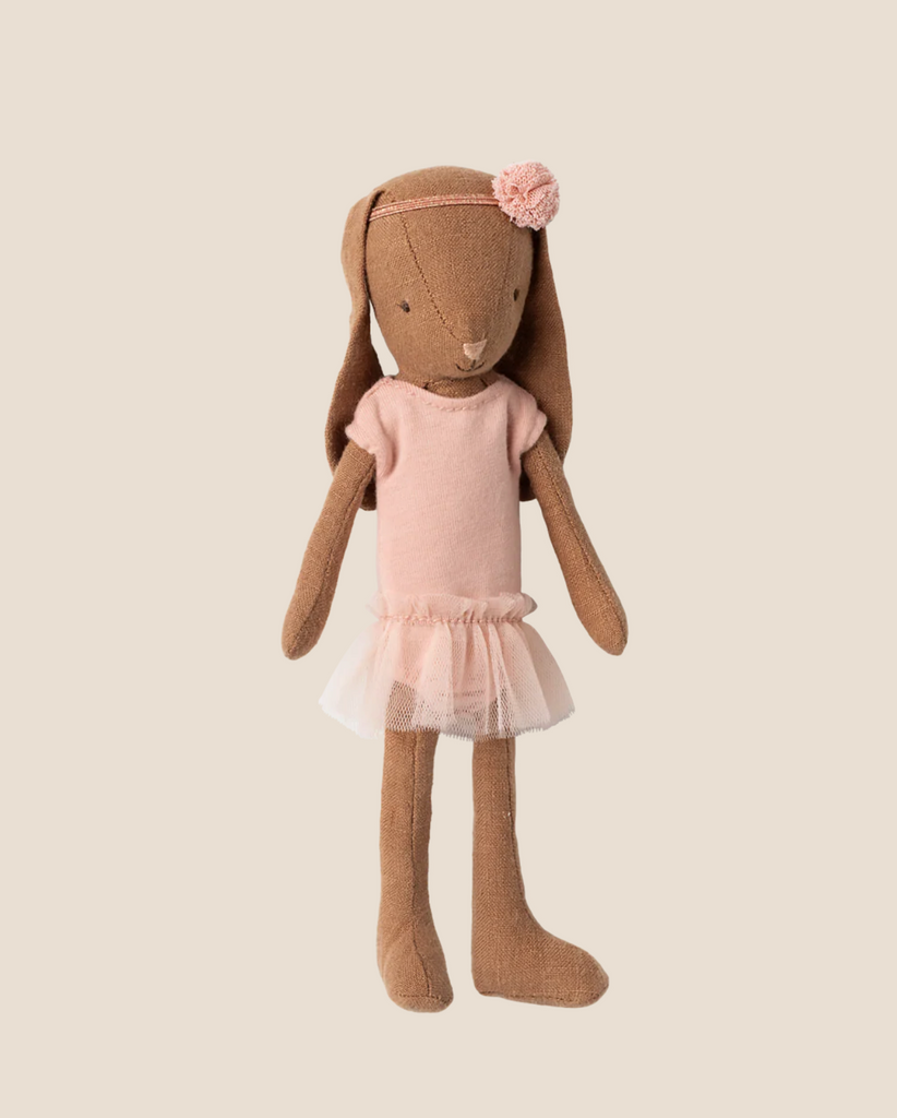 A Maileg Bunny, Size 1, in chocolate brown, wears a rose ballet suit and skirt. This adorable doll resembles a tiny ballerina and stands out beautifully against a plain beige background.