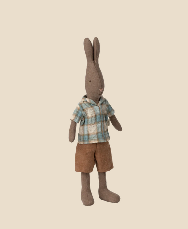 The Maileg Rabbit, Size 2 - Brown, stands upright in a blue and beige plaid shirt with brown shorts against a plain light beige background, crafted from natural fabrics for a charming touch.