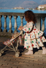 A young girl in a cream dress with colorful hearts pushes the Konges Sloejd Doll Stroller - Heart Kisses along a lakeside promenade bordered by stone railings, with mountains and water in the scenic background.