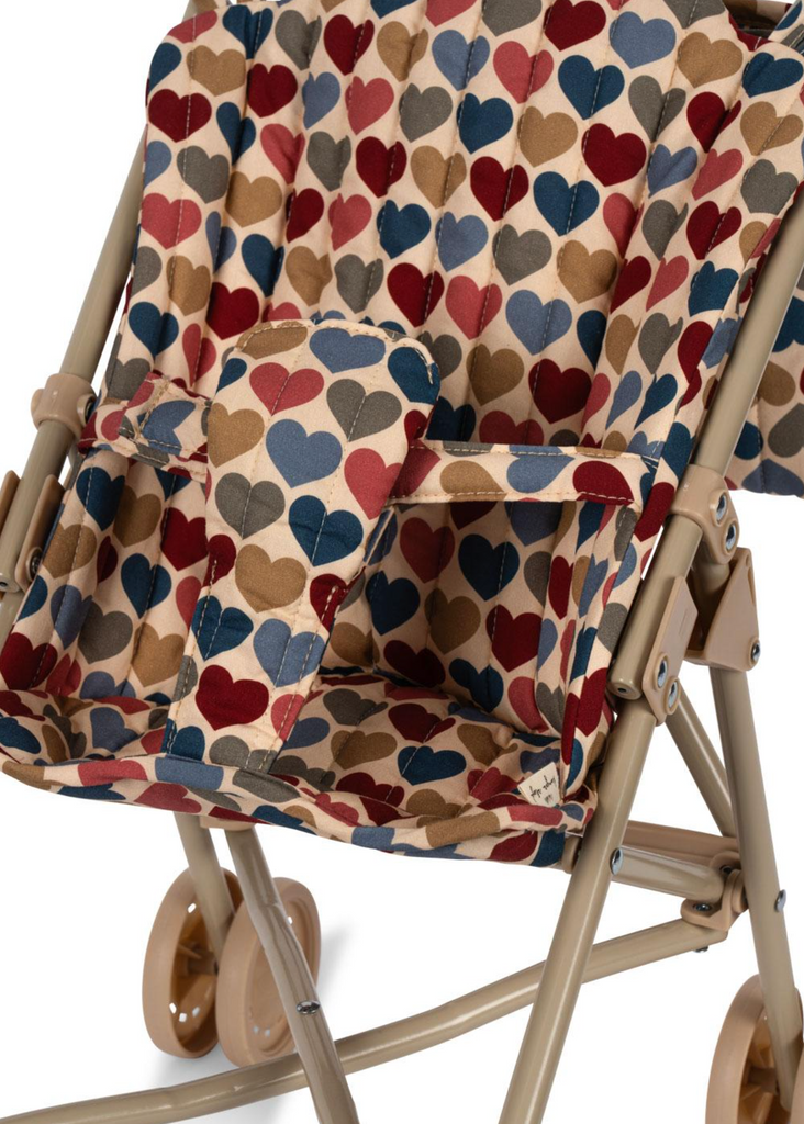 A close-up of the Konges Sloejd Doll Stroller - Heart Kisses shows its tan frame, double wheels, and armrests, with a fabric seat featuring a colorful heart pattern in red, blue, beige, and brown. The carriage bag is also partly visible.