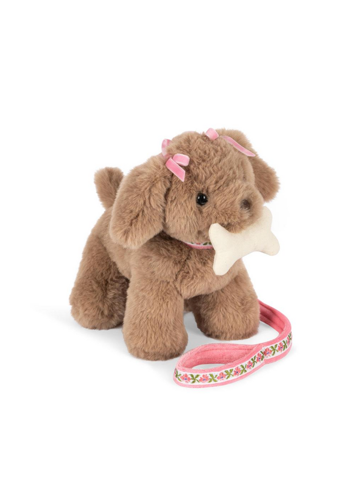 The Konges Sloejd Maude Puppy in Basket is a soft plush toy with pink bows on its ears, holding a gentle bone, and comes with a pink floral-patterned leash beside it.
