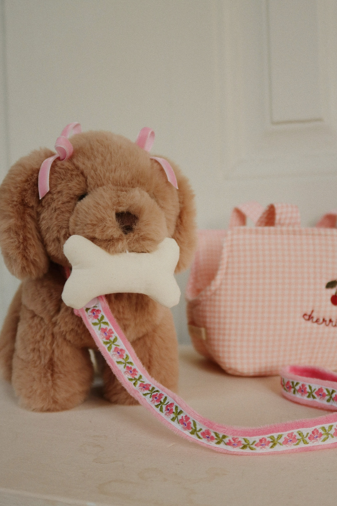 The Konges Sloejd Maude Puppy in Basket is a brown plush dog with pink ear bows, holding a white bone toy, featuring a pink floral leash, and comes with a pink gingham bag—perfect for carrying in its cute basket.