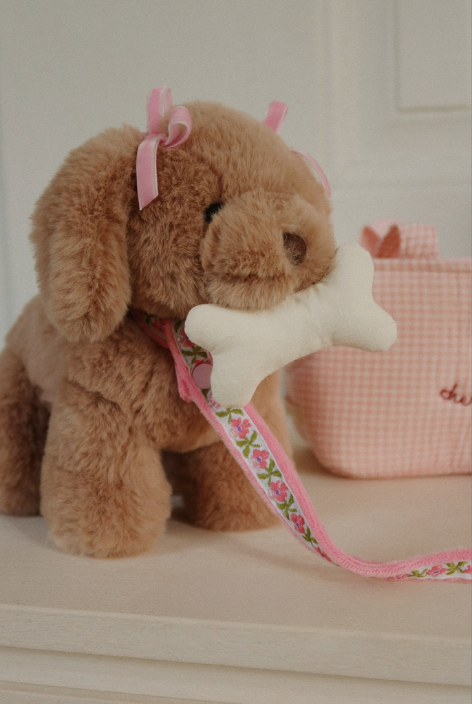 The Konges Sloejd Maude Puppy in Basket is a plush brown puppy with pink bows, a floral pink leash, and a white bone-shaped toy. A pink gingham bag is seen in the background.