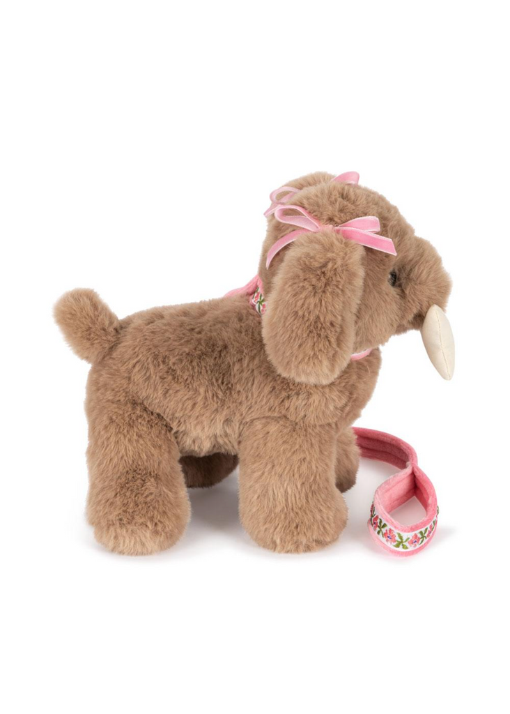 The Konges Sloejd Maude Puppy in Basket is a soft toy with floppy ears, pink ribbon bows, a floral leash, short tail, and plush fur. It stands on all fours facing left—perfect for cuddling or resting in its basket.