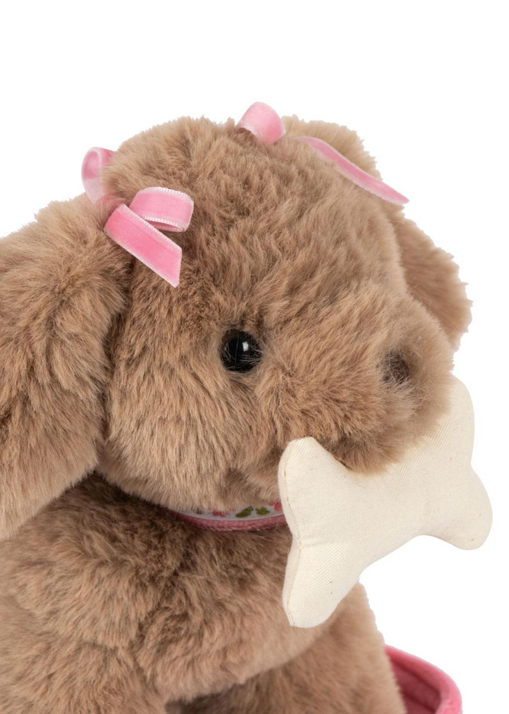 The Konges Sloejd Maude Puppy in Basket is a plush, CE certified brown dog toy with pink bows on its ears, holding a white bone-shaped toy—perfect for any dog basket.