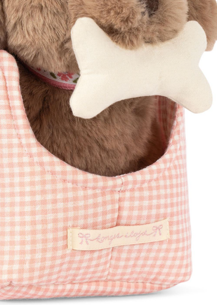 The Konges Sloejd Maude Puppy in Basket features a plush brown dog with a floral collar holding a white bone toy, seated in a pink and white checkered basket with an embroidered label.