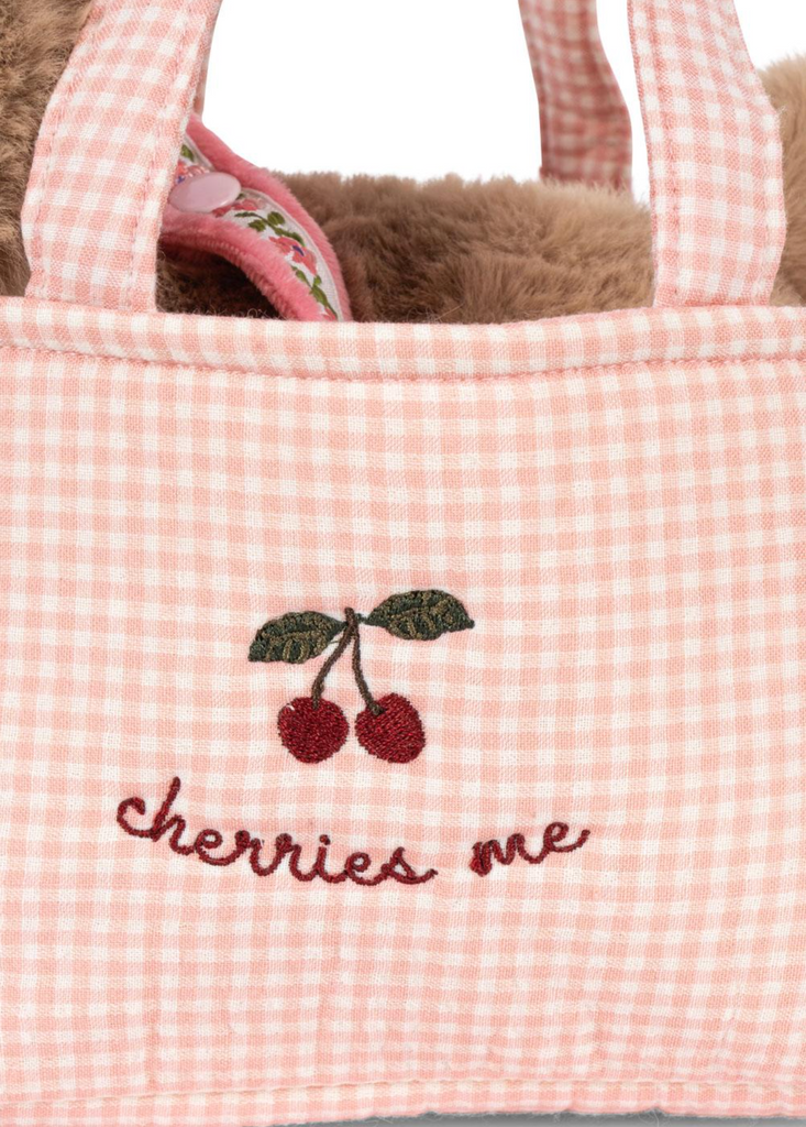 A close-up of the Konges Sloejd Maude Puppy in Basket: a pink and white gingham fabric bag with embroidered cherries and "cherries me" stitched in red, revealing a fuzzy brown lining, floral fabric, and a soft puppy toy inside.