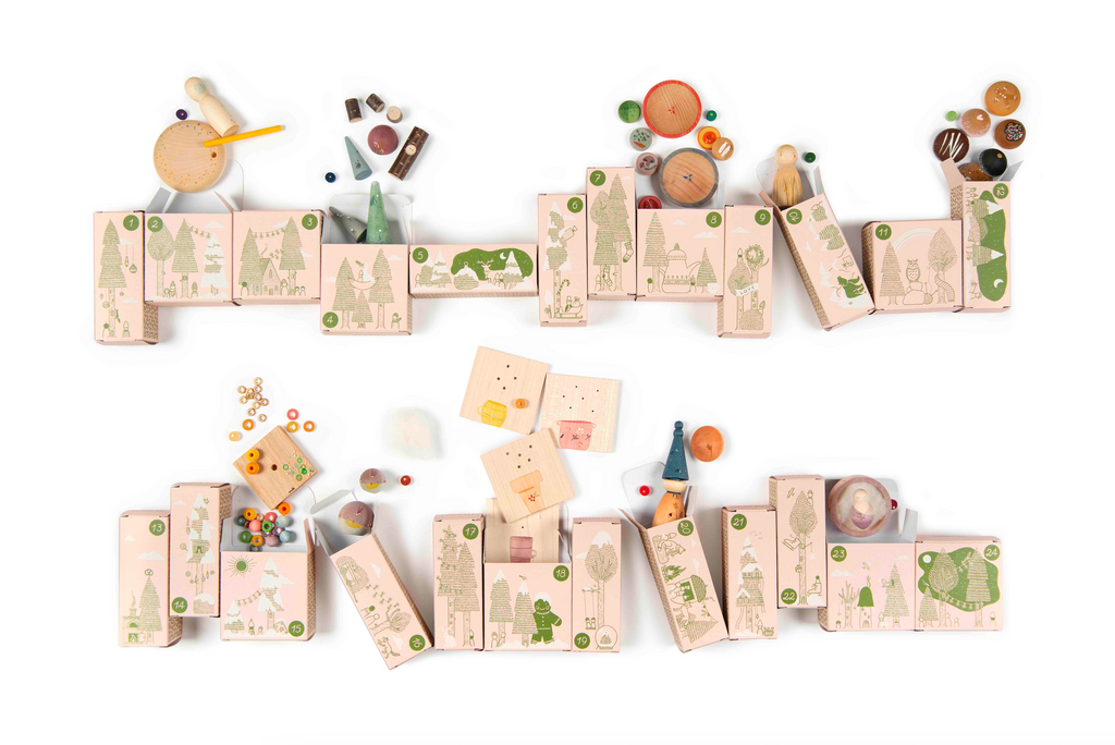 Wooden blocks with whimsical illustrations are arranged in two rows on a white surface, surrounded by colorful marbles, spinning tops, and toys from the Grapat Quiet Time Advent Calendar 2025. Two wooden figures and assorted pieces are also included.
