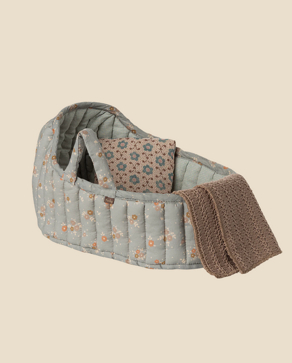 The Maileg Carry Cot, Large - Blue, is a soft, quilted baby doll carrier with a floral print, handles, matching pillow, and a textured brown knit blanket over the edge, ideal as a portable plush toy bed against a light beige background.