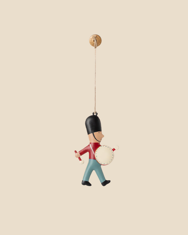 Add the Maileg Metal Ornament, Guard - Drum to your tree—a hand-painted hanging toy soldier in a black hat, red coat, and blue pants, marching with a drum. Ships in one week and is suspended by a string from a small round button.