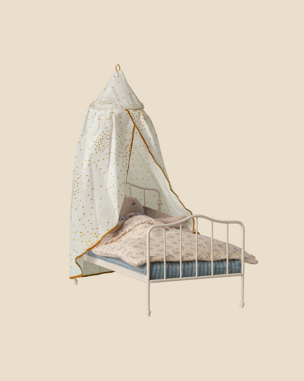 A white metal-framed bed with blue and pink bedding sits beneath the Pre-order: Maileg Bed Canopy, Miniature - Off-White (expected arrival by 04/15/26), adding a dreamy touch to the bedroom against a plain beige background.