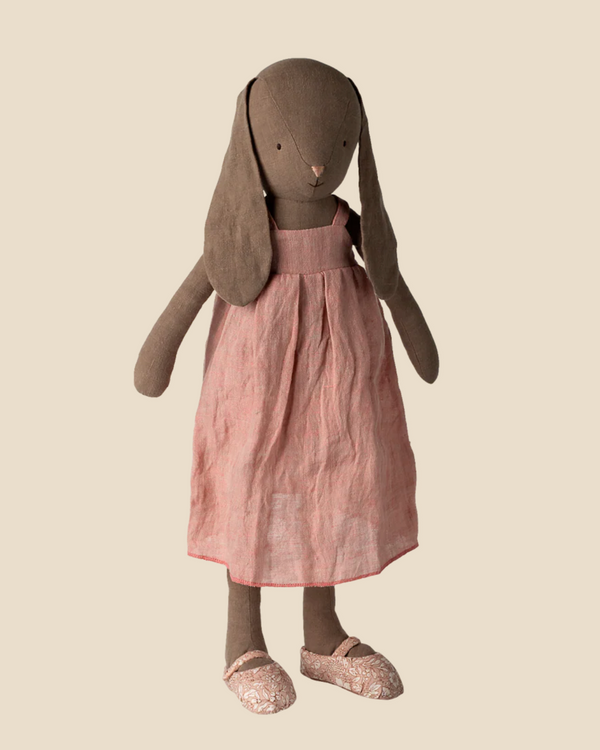 The Maileg Bunny, Size 4 in brown, wears a sleeveless pink dress and patterned shoes. This linen doll with floppy ears stands upright against a beige background. Pre-order now for expected arrival by 02/15/26.