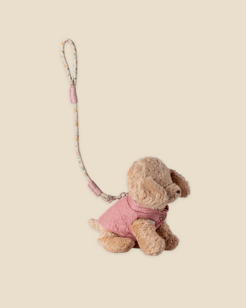 A small puppy plush toy models the Maileg Pet Supply, Harness and Leash - Rose (Pre-order, expected 03/01/26), attached to a patterned wrist strap with a metal clasp and cute accessories, set against a beige background.