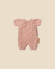 The Maileg Little Lambs Kitchen Set, featuring a small pink knitted baby romper with short sleeves and legs, is laid flat on a beige background with a fabric tag attached—perfect for enhancing your Maileg kitchen display.