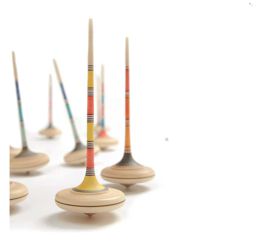 Several tall, colorful spinning tops—including the Mader Arabella Spinning Top in Purple—stand upright on a white surface in a scattered arrangement, some in sharp focus and others blurred, highlighting the toy's safety features.