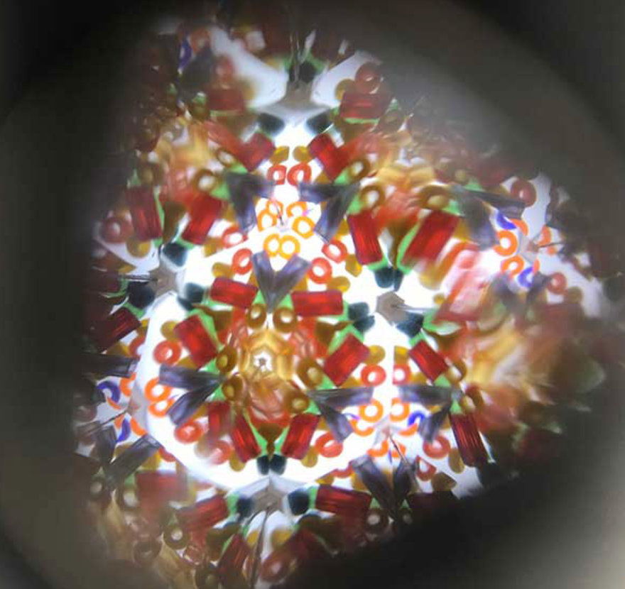 Peer through the Mader Wooden Kaleidoscope - Dark Blue to see a classic toy’s symmetrical, colorful patterns—red, green, orange, blue, and yellow shapes on a white background—all framed by a dark circular border.