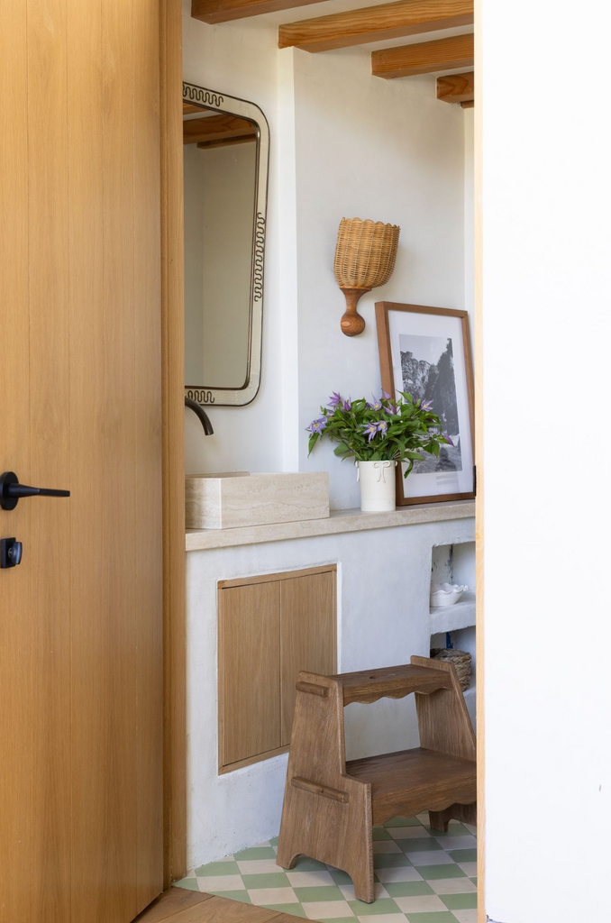 A cozy entryway with a wooden door, vintage mirror, small countertop displaying a framed photo, vase of flowers, woven wall basket, and the Margot Step Stool on a green-and-white checkered tile floor.
