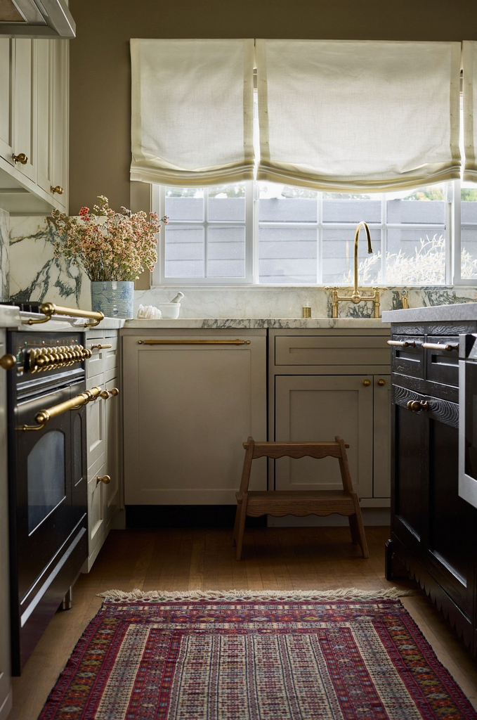 A cozy kitchen with white cabinets, marble countertops, gold hardware, a vintage rug, flowers in a vase, sunlight streaming through white Roman shades above the sink, and a Margot Step Stool made of solid oak.