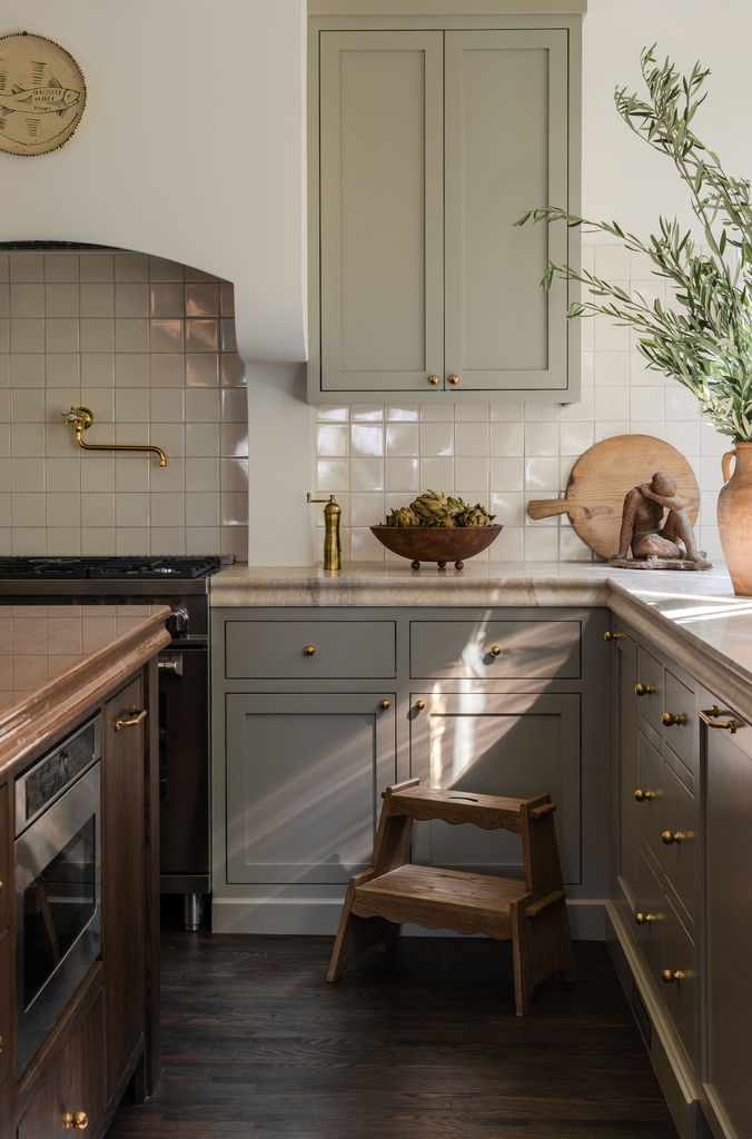 A cozy kitchen features gray cabinets, brass hardware, a tiled backsplash, wooden accents like cutting boards and a vase with greenery, and the Margot Step Stool in solid oak. Sunlight streams onto the countertop.
