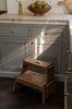 The Margot Step Stool stands on a dark wood floor in front of gray kitchen cabinets with brass knobs. Sunlight streams in, casting bright streaks and shadows across the cabinets and the Margot Step Stool.