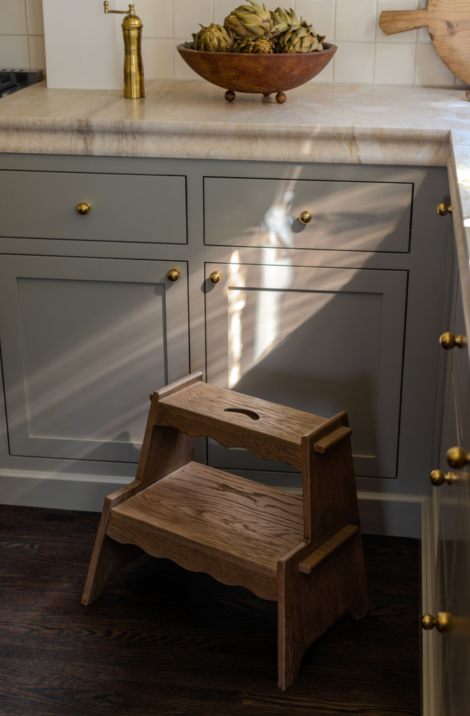 The Margot Step Stool stands on a dark wood floor in front of gray kitchen cabinets with brass knobs. Sunlight streams in, casting bright streaks and shadows across the cabinets and the Margot Step Stool.