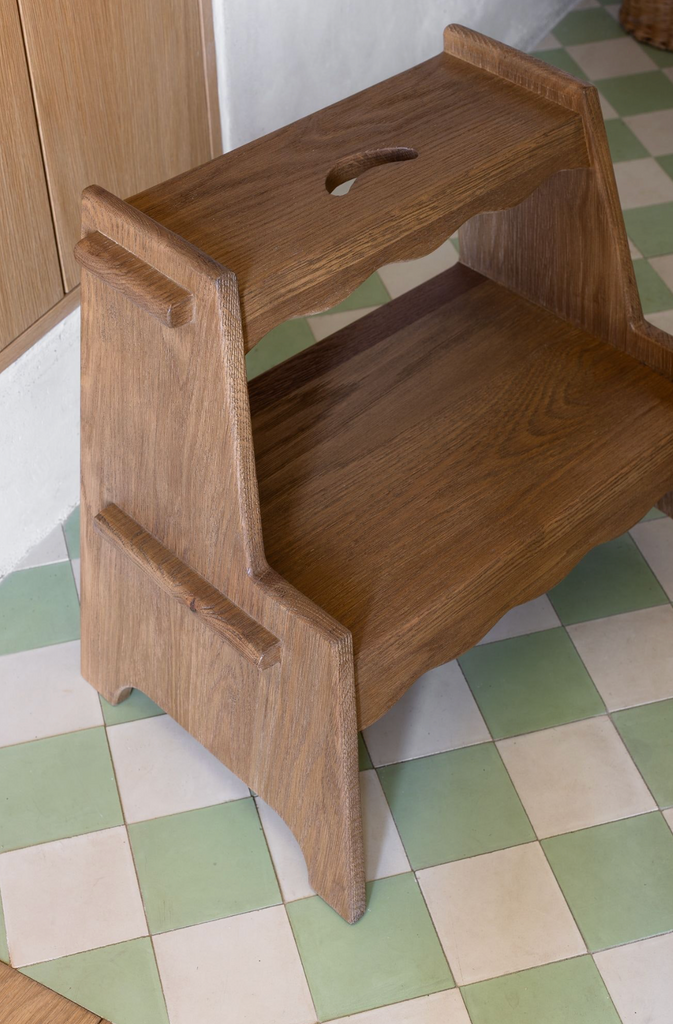 The Margot Step Stool, made of solid oak with two steps and a handle cutout on top—ideal for kids—is placed on a green and white checkered tile floor.