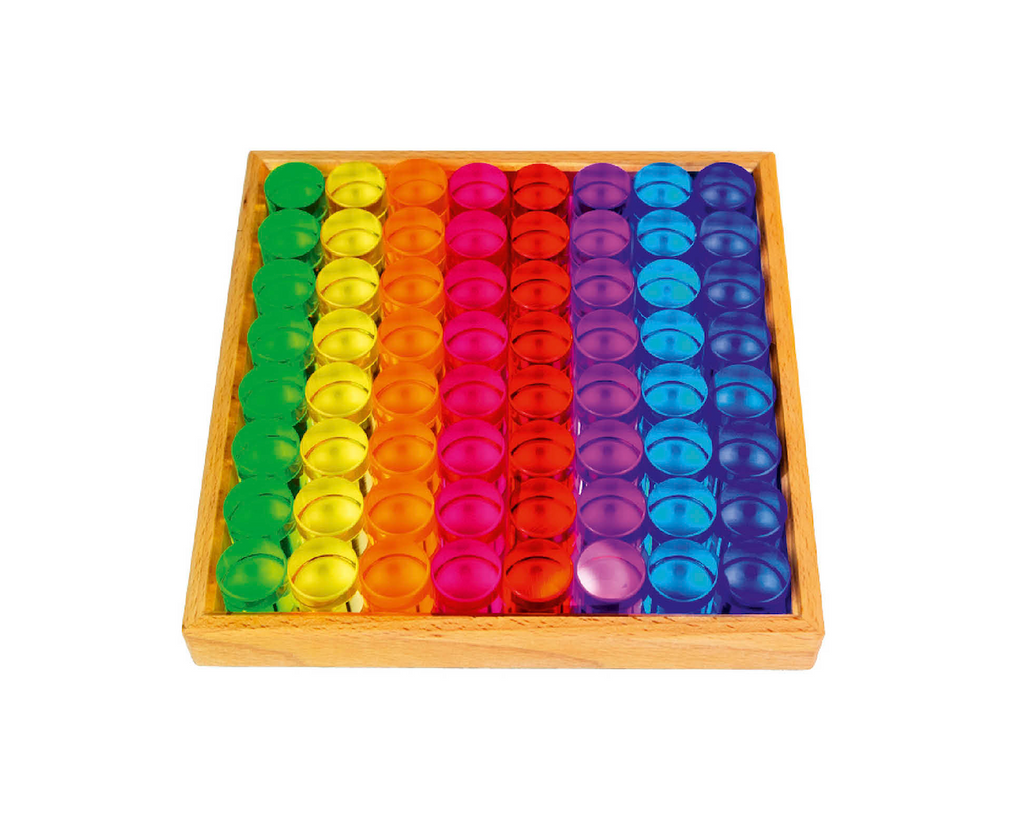 A wooden tray holds rows of Bauspiel Lucent Cylinders—round, colorful translucent tokens—arranged in a rainbow pattern with green, yellow, orange, red, pink, purple, and blue sections in neat lines.