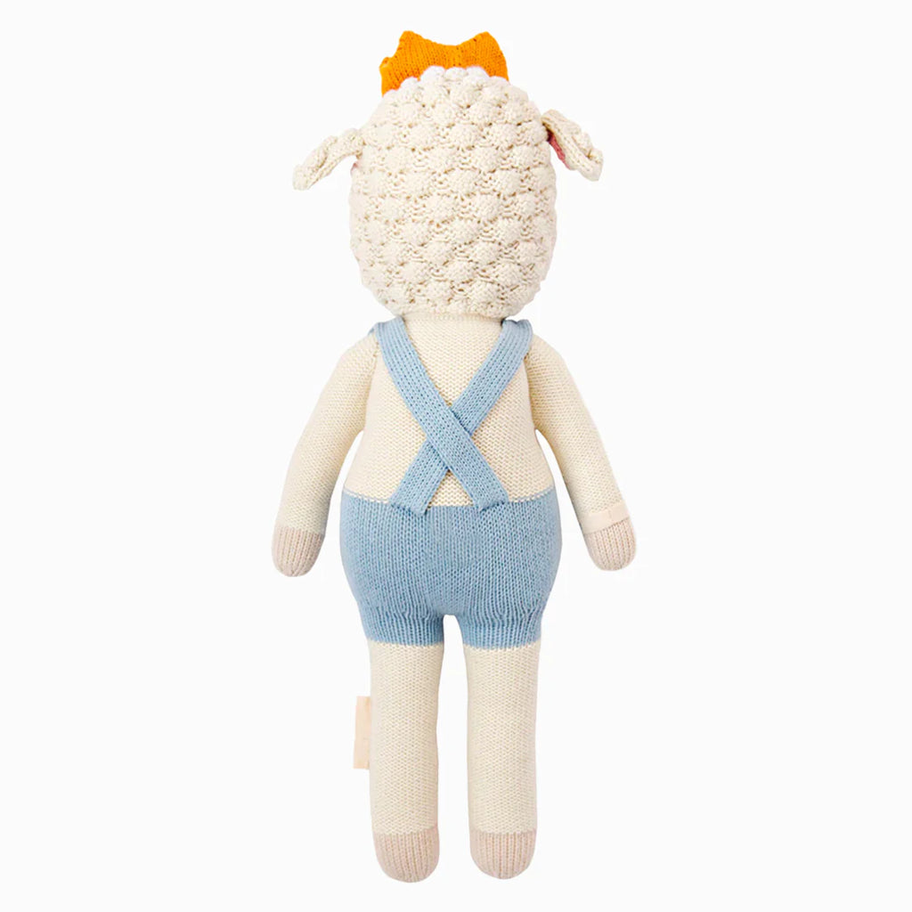 Cuddle + Kind Sebastian the Lamb is a hand-knit doll in light blue crisscross overalls and an orange crown, seen from the back. Made using sustainable fair trade practices. Background is plain white.