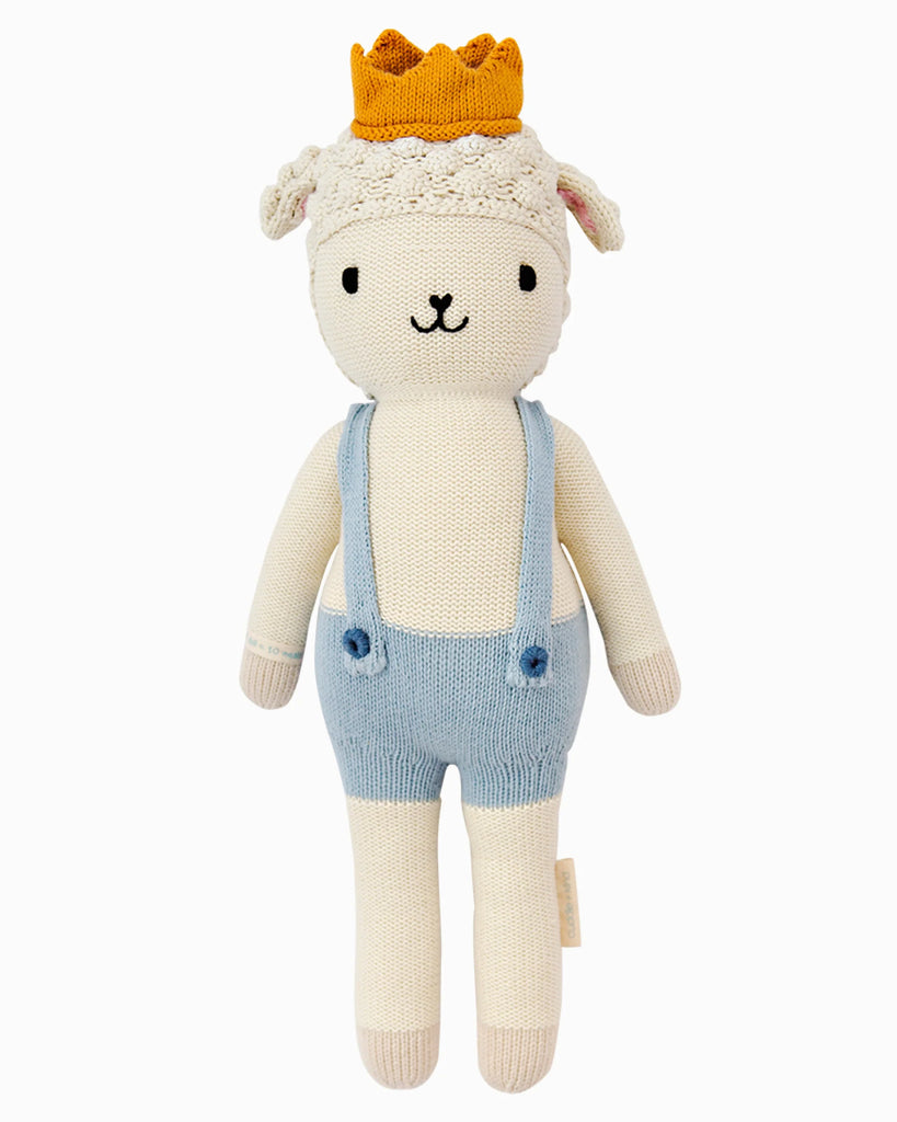 Cuddle + Kind Sebastian the Lamb is a hand-knit doll with a neutral expression, yellow crown, light blue suspenders, and white shorts—crafted with sustainable, fair trade practices.