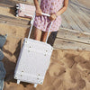 A person wearing a pink polka-dotted dress stands on a sandy surface, holding the handle of a Olli Ella See-Ya Suitcase - Leafed Mushroom. Only the lower half of the person is visible.