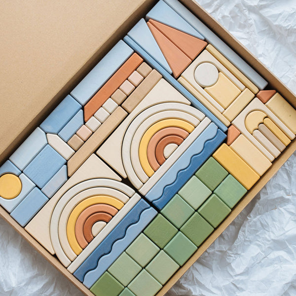 A Meshka & Friends Block Set, handcrafted in Serbia, arranged neatly in a box and featuring various shapes and patterns in shades of blue, yellow, green, and natural wood.