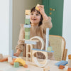 Young girl joyfully playing with the Meshka & Friends Block Set and building a tall structure on a table, in a bright room.