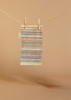 A small, multicolored woven mat with fringe hangs from a clothesline by two wooden clothespins against a beige background—an adorable accessory for your Maileg Fully Furnished SuitCasa.