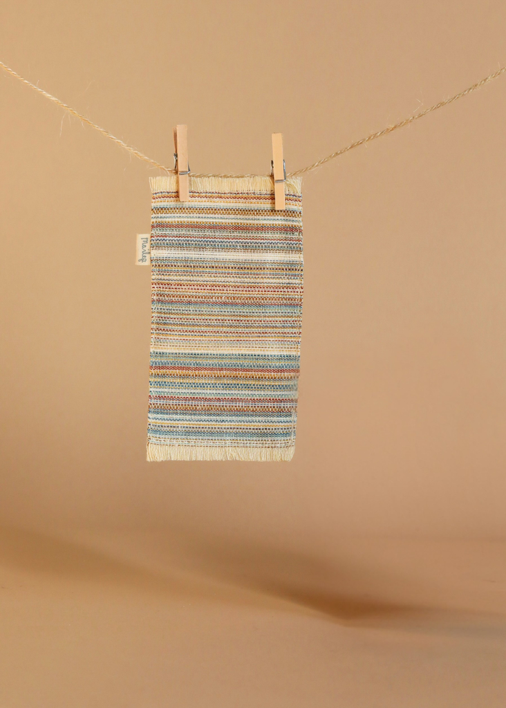 A small, multicolored woven mat with fringe hangs from a clothesline by two wooden clothespins against a beige background—an adorable accessory for your Maileg Fully Furnished SuitCasa.