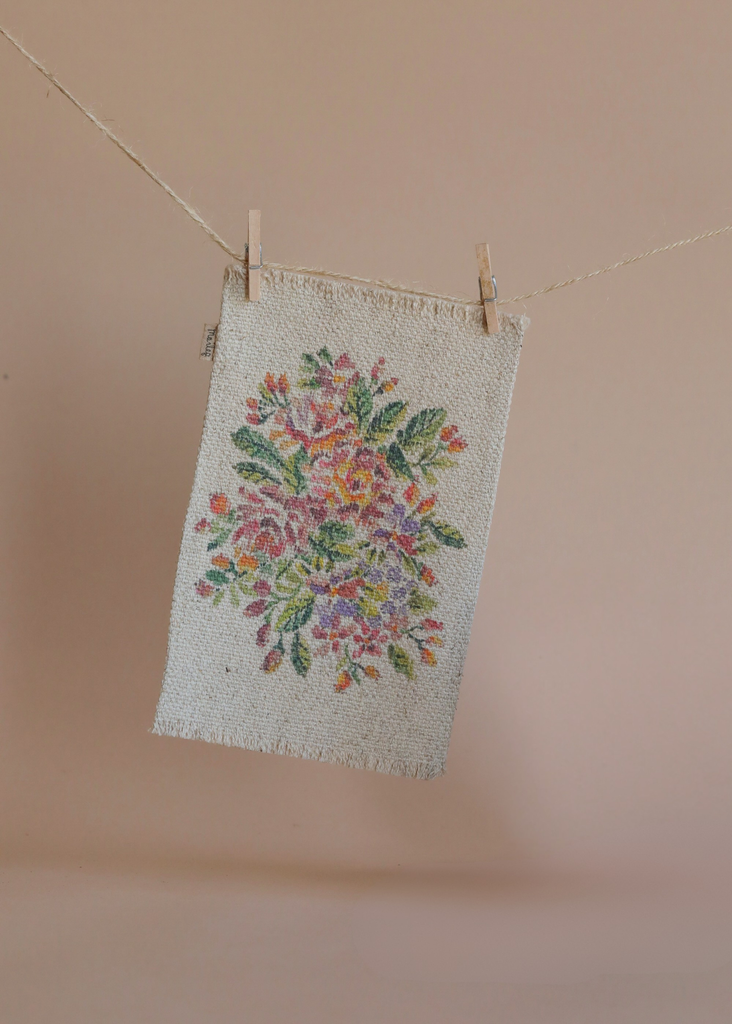 A piece of fabric from the Maileg Family Christmas Set, embroidered with a colorful floral pattern, hangs from a clothesline with two wooden clothespins against a plain beige background.