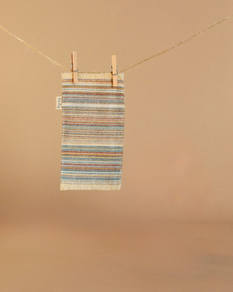 A small, striped woven rug in earth tones hangs from a clothesline with wooden clothespins against a beige background, perfectly complementing the Maileg Little Living Room Set for a cozy touch.