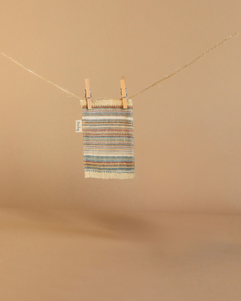 A small multicolored woven textile hangs on a string with two wooden clothespins against a beige background, resembling cozy miniature accessories from the Maileg Cozy Green Room Set.