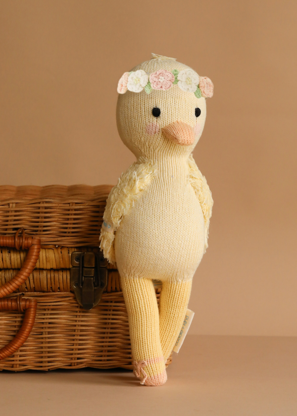Cuddle + Kind Flora the Duckling - Mauve, a soft knit cotton doll with a floral crown, leans against a wicker basket on a plain beige background.