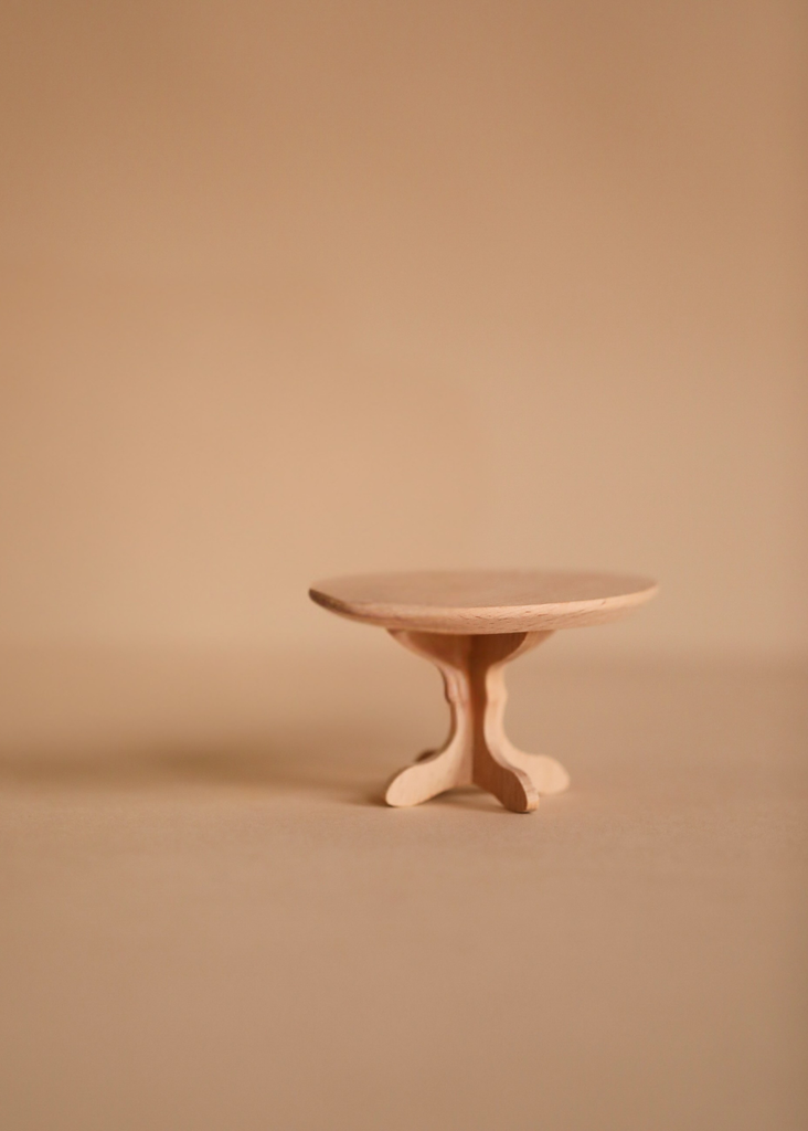 The Maileg Fully Furnished SuitCasa, featuring a small, round wooden table with three curved legs, stands on a beige surface against a matching background—ideal for displaying Maileg kitchen accessories or mouse house décor.