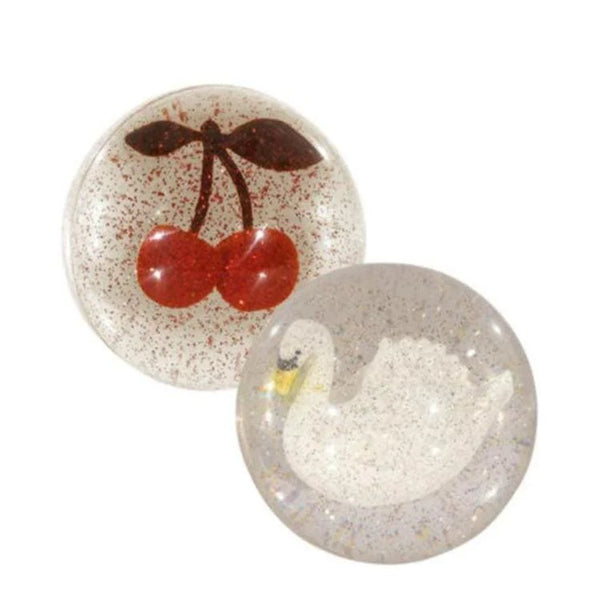 The Konges Sloejd Glitter Bouncing Ball Set includes two clear resin balls: one with red cherries and green stems, the other with a white swan, both featuring a glittery background for a sparkling effect.