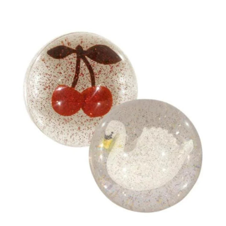 The Konges Sloejd Glitter Bouncing Ball Set includes two clear resin balls: one with red cherries and green stems, the other with a white swan, both featuring a glittery background for a sparkling effect.