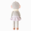 A fair trade toy, Cuddle + Kind Skylar The Penguin is a handmade knitted stuffed animal resembling a penguin, shown from the back, wearing a light purple skirt, cream-colored scarf, and pink earmuffs, with brown feet and gray fuzzy arms.